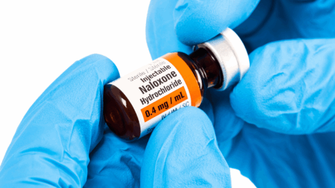 Close-up of gloved hands holding a vial of injectable naloxone (0.4 mg/mL), used to reverse opioid overdoses