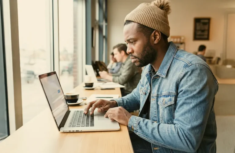 Man in beanie and denim jacket researching addiction relapse and overdose risks on laptop in a caf&eacute;