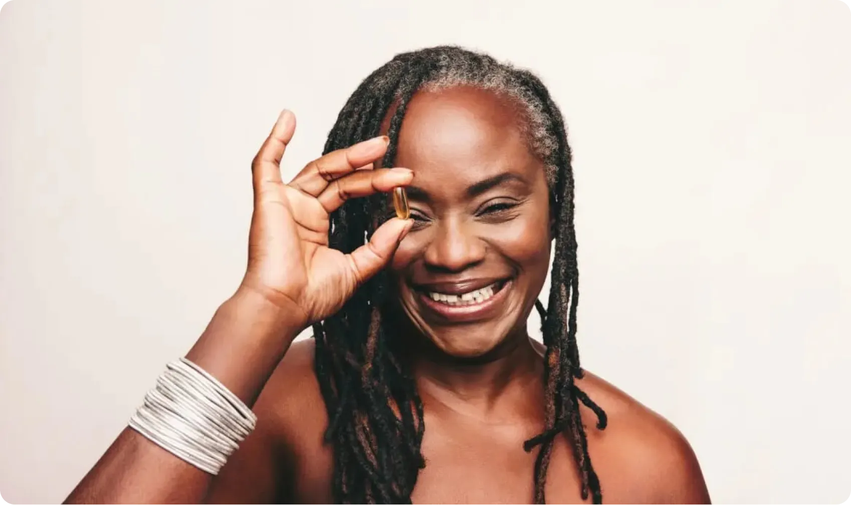 Cheerful woman with dreadlocks smiling while holding a small supplement capsule near her face against a light background