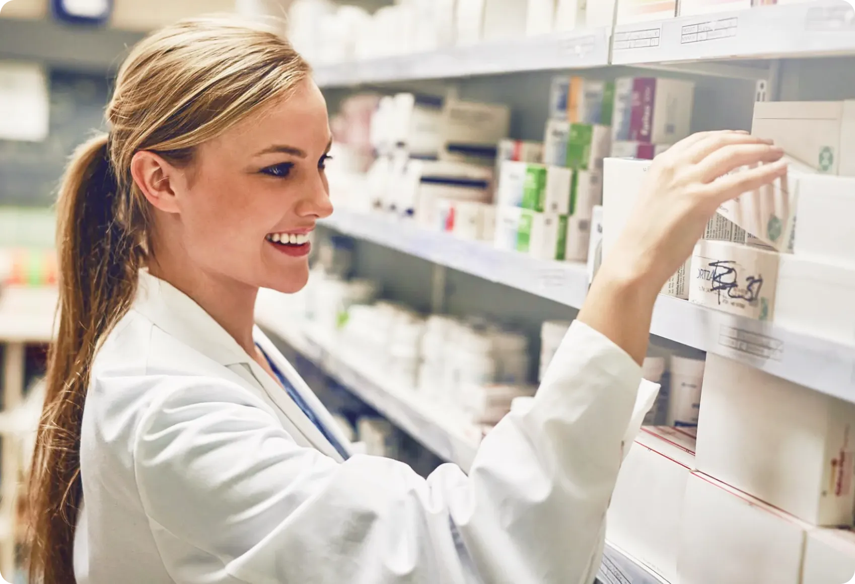 Smiling female pharmacist in white coat retrieving medication from a well-stocked pharmacy shelf