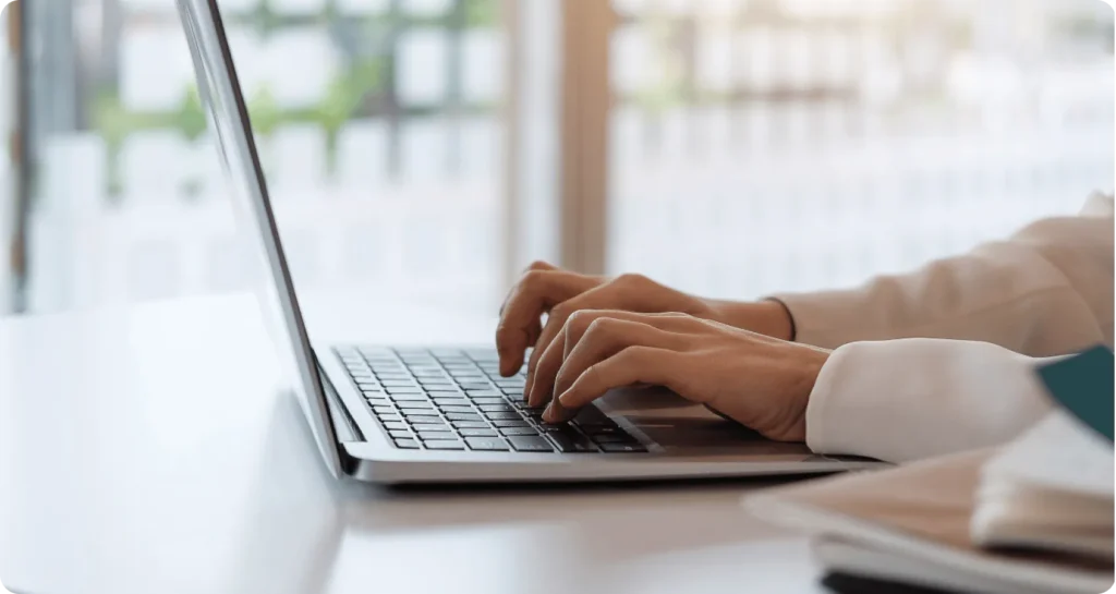 Person in a white coat typing on a laptop at a bright, modern desk in a professional setting