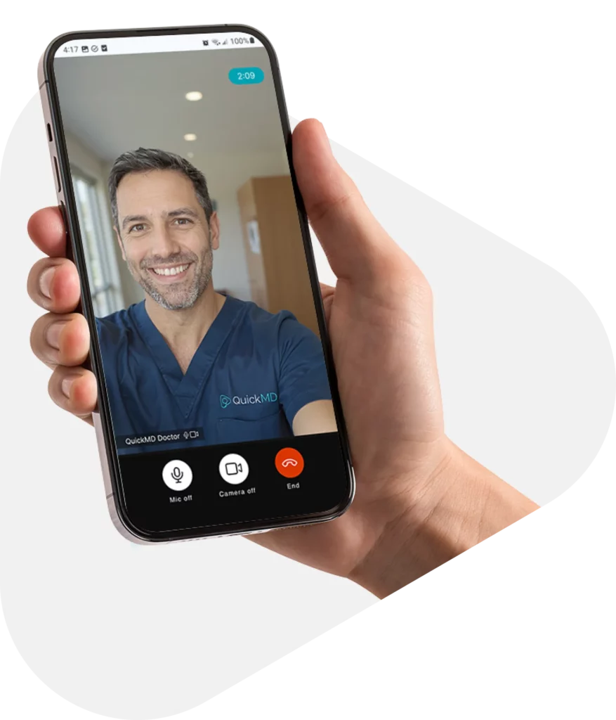 Hand holding a smartphone during a QuickMD telehealth video call with a smiling doctor in blue scrubs