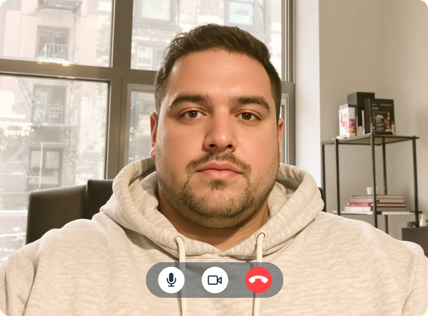 Man in a beige hoodie on a telehealth call, sitting in a modern apartment with city view windows behind him.