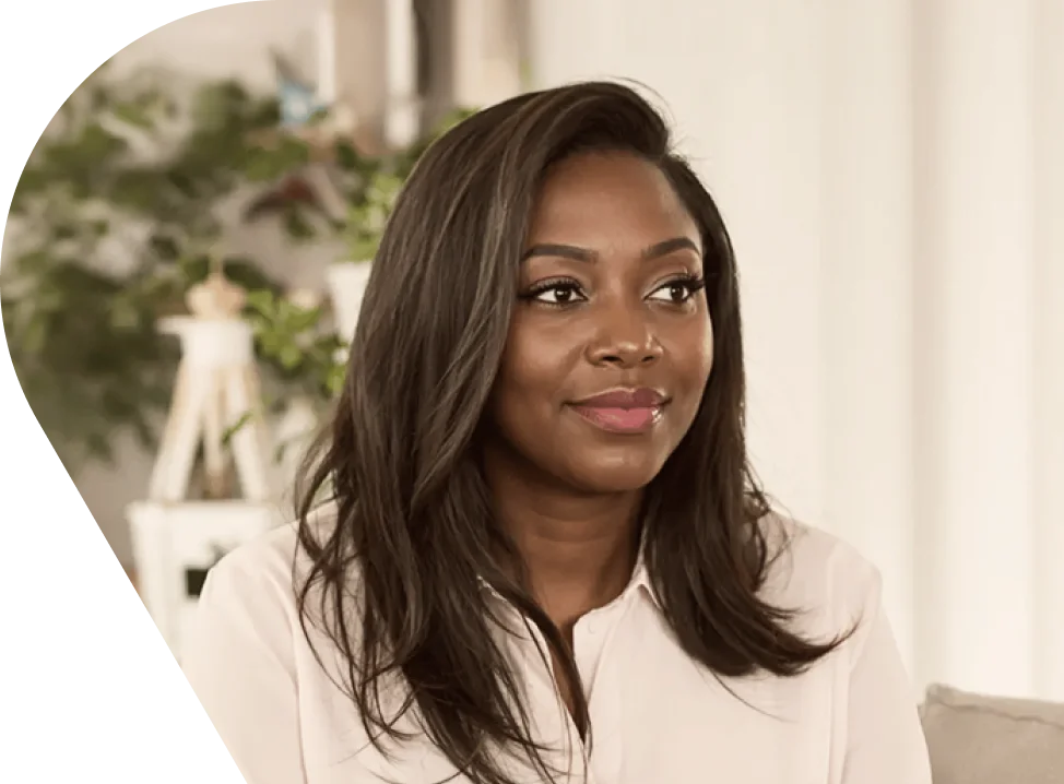 Smiling young Black woman with long dark hair wearing a white blouse, seated in a bright, plant-filled living room