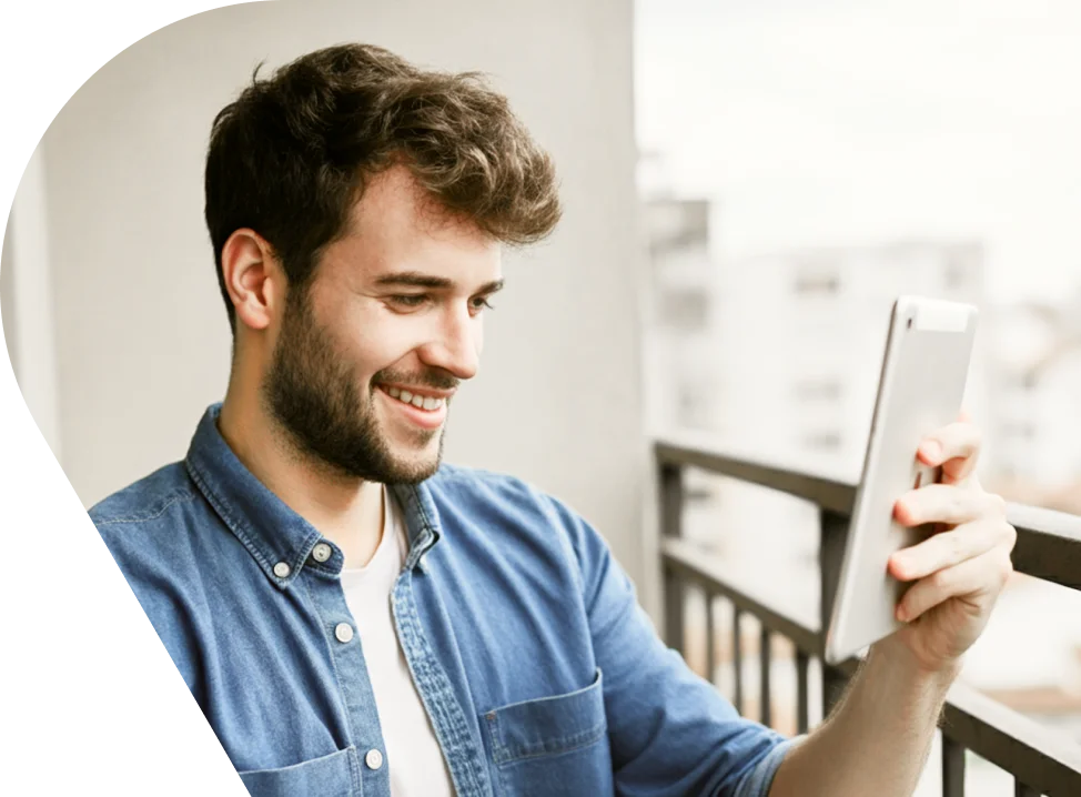Smiling young bearded man in denim shirt holding a tablet device outdoors on a balcony
