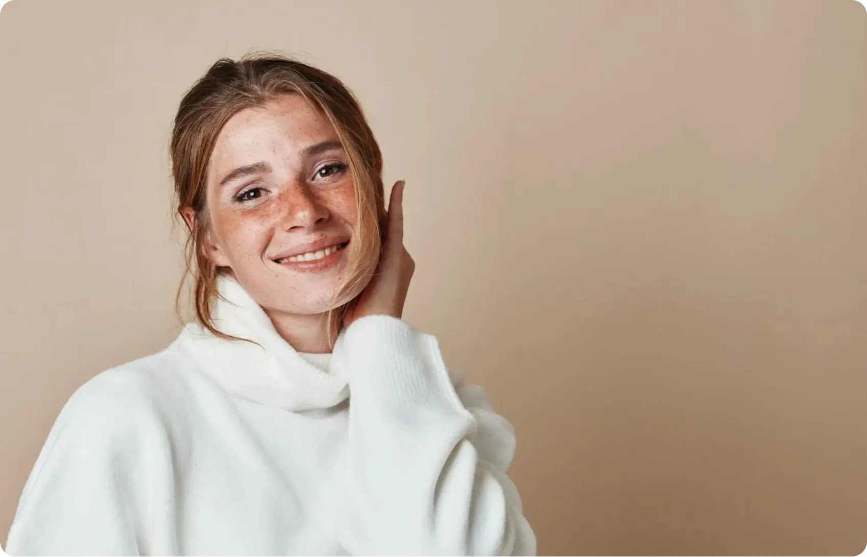 Smiling young woman with freckles wearing a white turtleneck sweater, touching her face against a beige background
