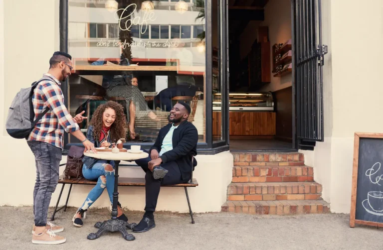 Group of friends laughing and talking outside a caf&eacute;, representing building a supportive recovery community with help from telehealth care like QuickMD.