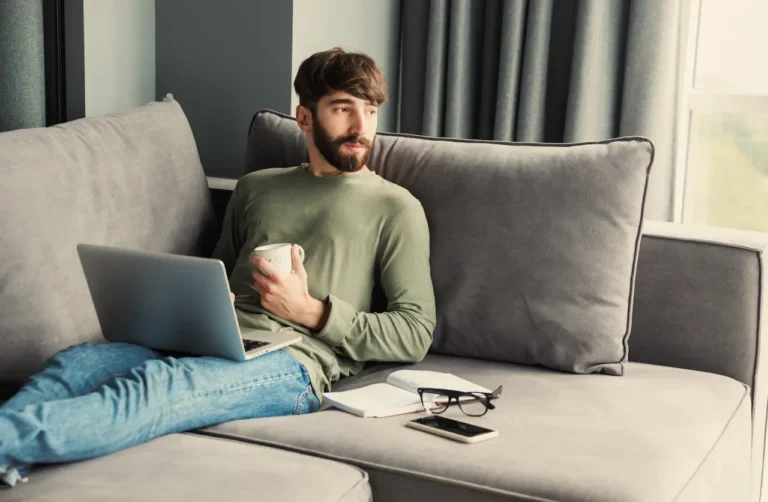 Man sitting on a couch with a laptop and coffee, appearing thoughtful, representing mental health support during Suboxone treatment through telehealth care like QuickMD.