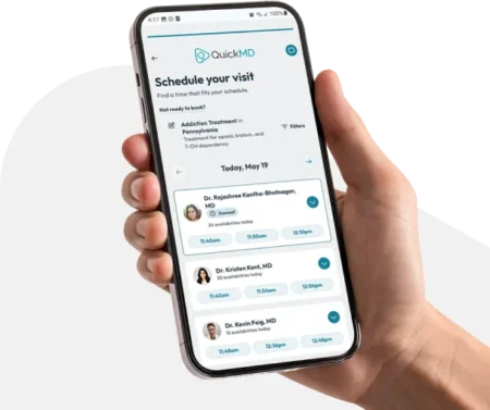 Hand holding a smartphone displaying the QuickMD app's "Schedule Your Visit" screen with available doctors and time slots.