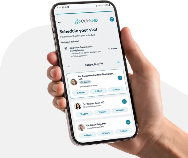 Hand holding a smartphone displaying the QuickMD app's "Schedule Your Visit" screen with available doctors and time slots.