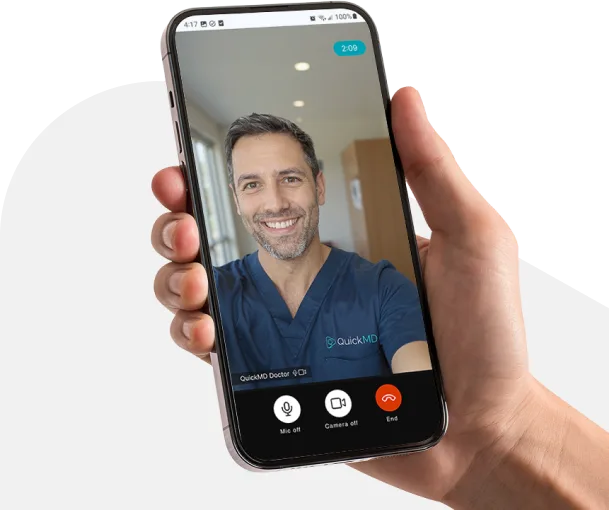 Hand holding smartphone showing a QuickMD doctor video call consultation with a smiling male physician in scrubs