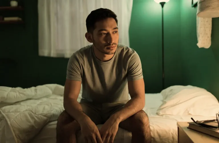 man-sitting-on-bed-looking-distressed-in-dimly-lit-bedroom-depicting-kratom-withdrawal-symptoms-and-discomfort