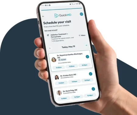 Hand holding a smartphone displaying the QuickMD app's "Schedule Your Visit" screen with available clinicians and time slots.