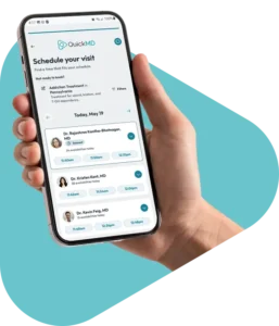 Hand holding a smartphone displaying the QuickMD app's "Schedule Your Visit" screen with available doctor appointment times.