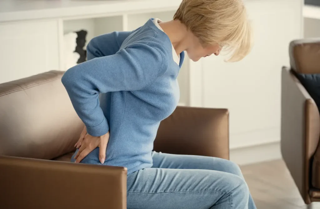 Middle-aged woman sitting in a chair holding her lower back, illustrating back pain associated with menopause and osteoporosis.