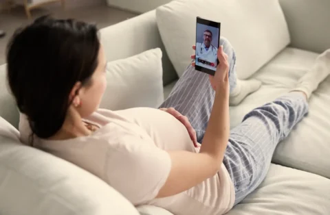 Pregnant woman lying on a couch at home during a telehealth video call with a doctor on her smartphone.