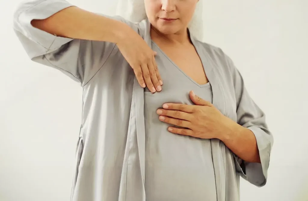 Middle-aged woman wearing a gray robe gently examining her breast with one hand on her chest and the other raised, illustrating breast changes during menopause.