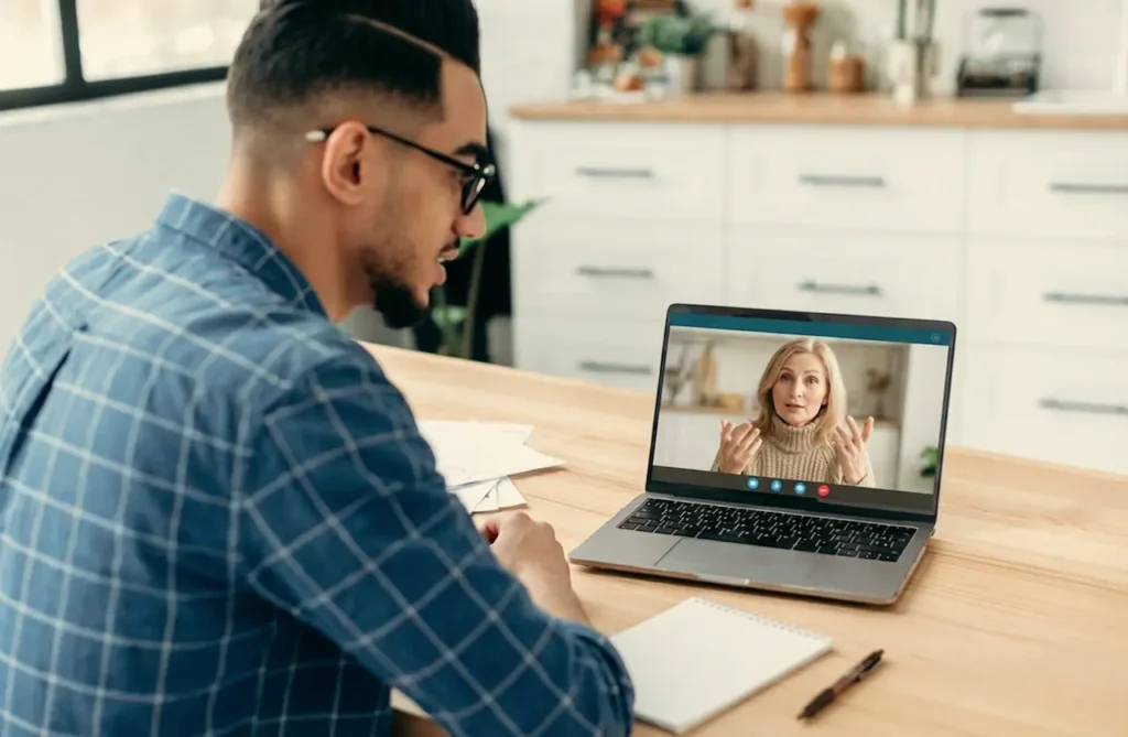 Man sitting at table at home during a telehealth video appointment on his laptop, speaking with a female healthcare provider on screen.