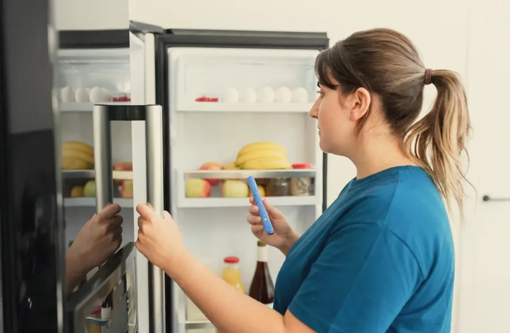 Woman standing in front of an open refrigerator holding an Ozempic injection pen, with food items visible inside the fridge.