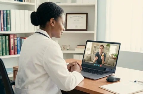 Doctor in a white coat sitting at a desk in a medical office, conducting a telehealth video call on a laptop with a male patient holding a prescription pill bottle.
