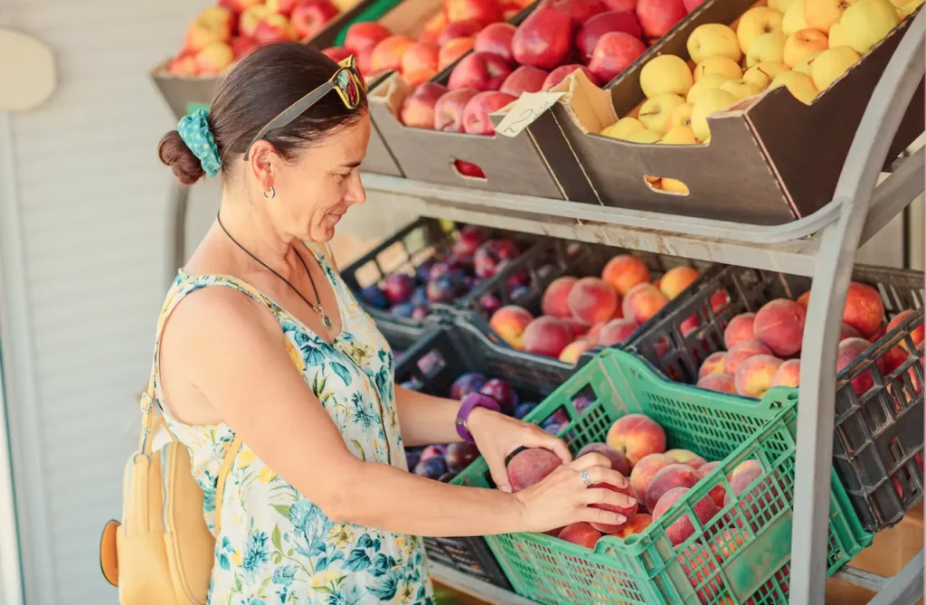 Woman selecting fresh peaches from a produce display at a market, with crates of apples and stone fruit on shelves behind her.